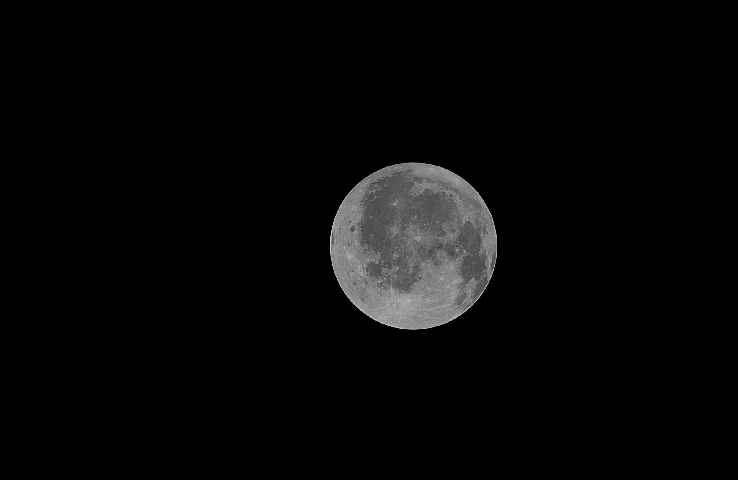 A near-full Moon photographed in high detail against a pure black sky. The lunar surface is rendered in crisp greyscale, with dark maria and lighter highland regions clearly visible across the face. The Moon is positioned slightly right of centre, with no other elements in the frame. Stark and isolated.