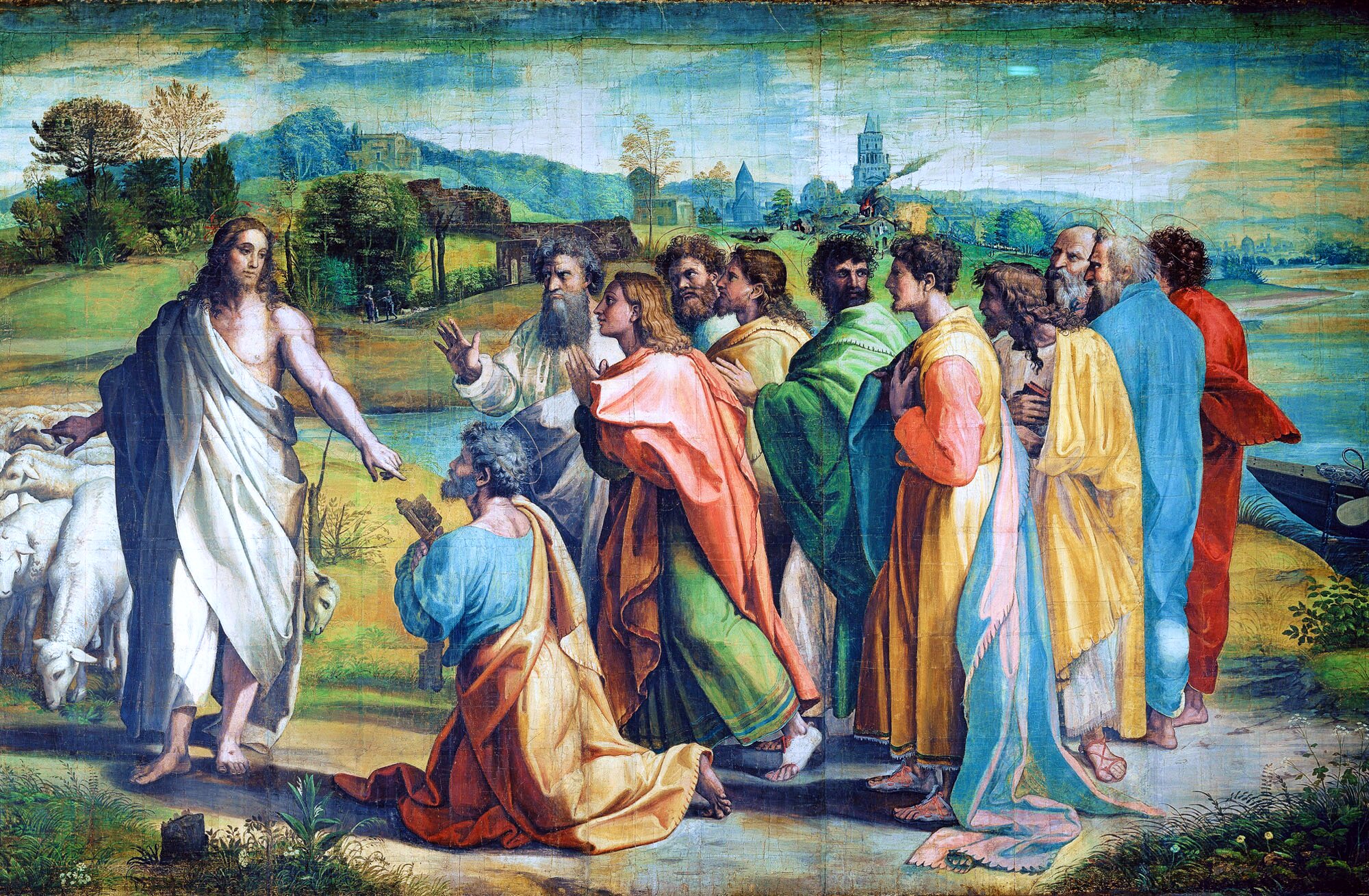 A robed Christ stands at left, gesturing toward a group of apostles in a lush pastoral landscape. At centre, an older bearded man—Peter—kneels in supplication before him, arms raised. Behind Peter, a crowd of apostles in vibrant draped robes of coral, green, gold, blue, and red look on with varying expressions of attention and concern. A flock of white sheep grazes at left. In the background, rolling green hills, scattered trees, and the towers of a distant village recede under a pale blue sky. The painting surface shows visible cracking consistent with age.
