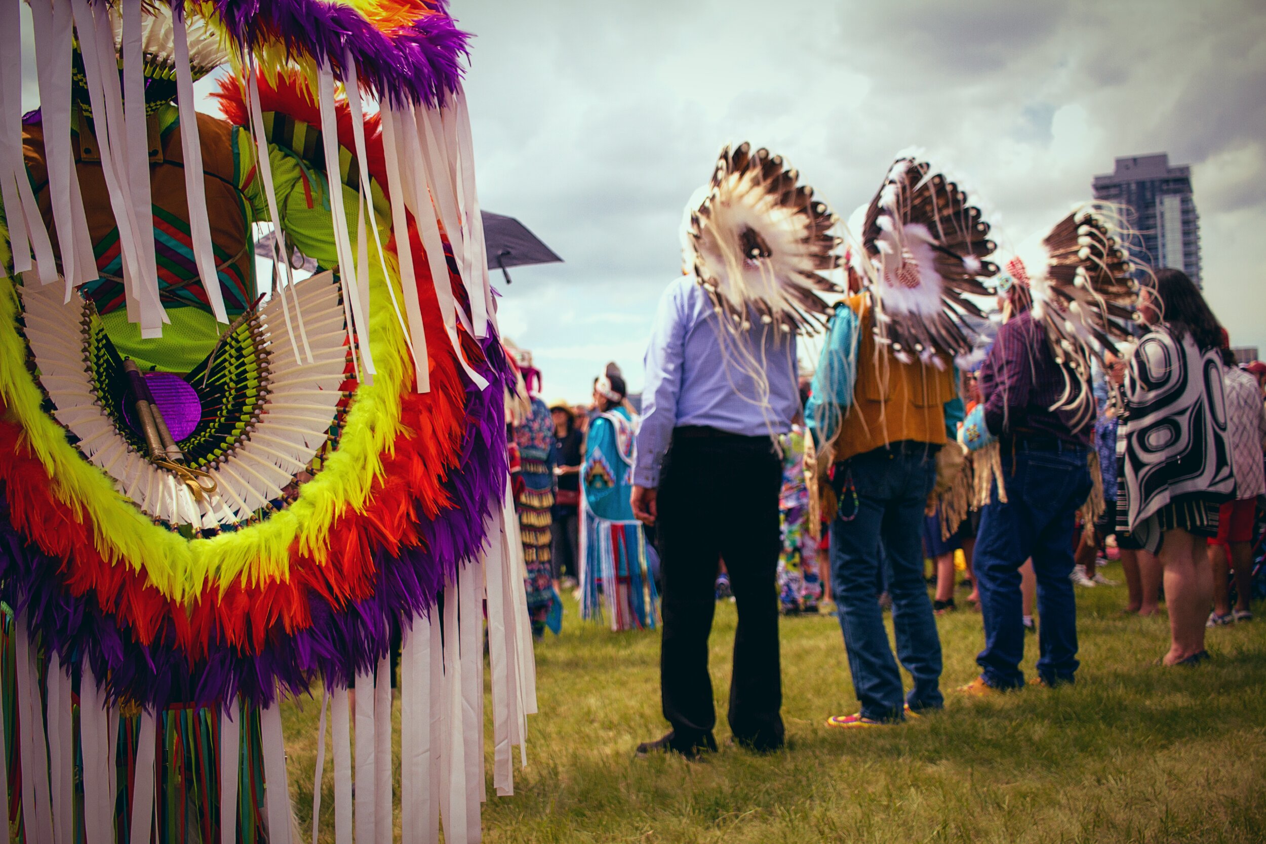 An outdoor powwow or Indigenous ceremonial gathering set against an overcast urban skyline. The left foreground is filled with a large, strikingly colourful regalia bustle. Concentric rings of neon yellow, red, orange, and purple feathers with long white ribbon fringe. In the mid-ground, several figures stand on a grassy field; two wear large traditional eagle feather headdresses that face away from the camera, their faces obscured. One person wears a Northwest Coast-style button blanket or cape featuring black-and-white formline design. The gathering includes people in both traditional regalia and everyday clothing. A highrise tower is visible through the cloudy sky in the distance, situating this cultural gathering within a contemporary urban landscape.