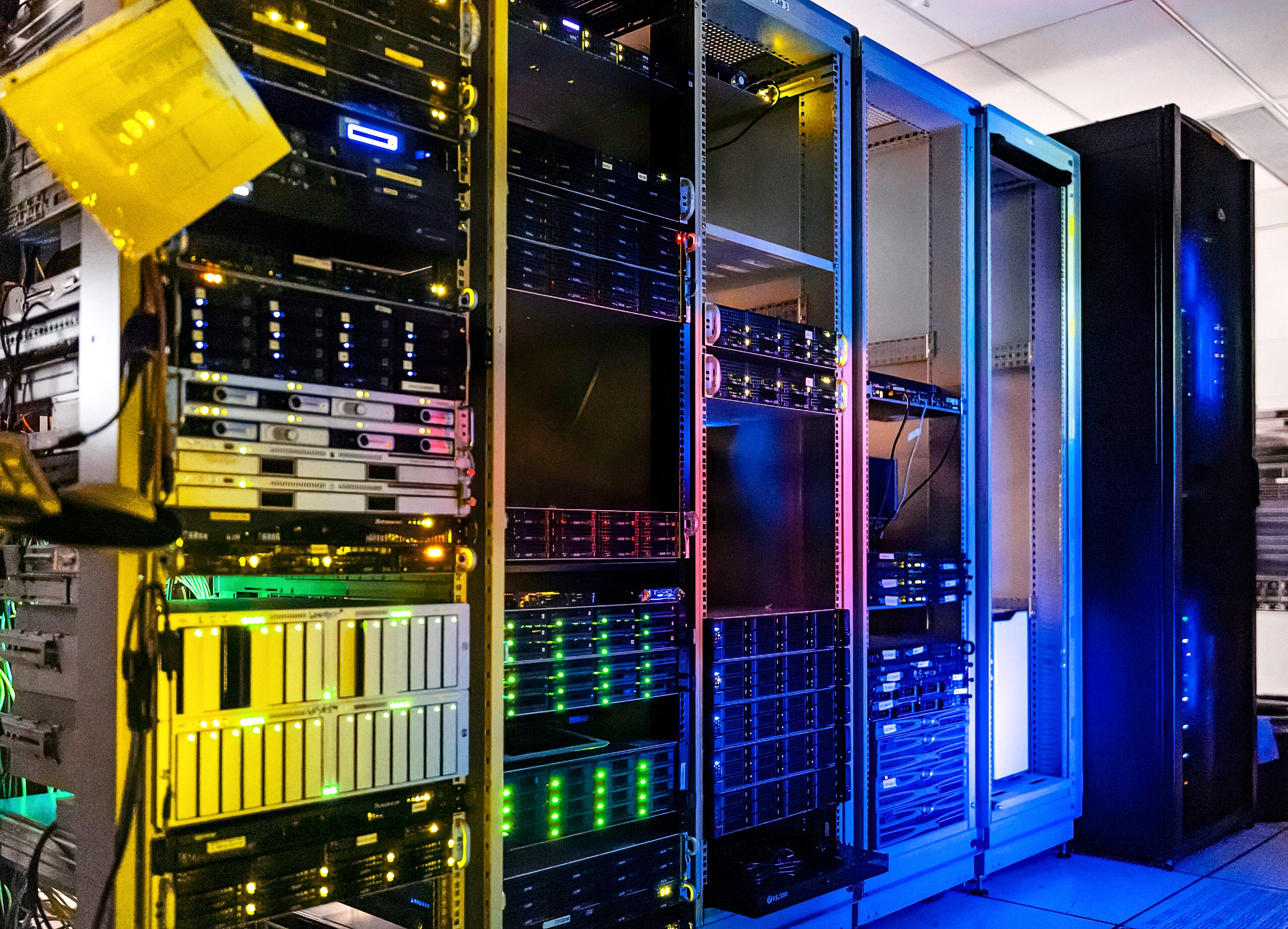 A server room with multiple open rack cabinets filled with densely packed hardware, illuminated by dramatic coloured lighting: yellow on the left, pink and green in the centre, and blue on the right. Status indicator LEDs in amber, green, and red dot the equipment.