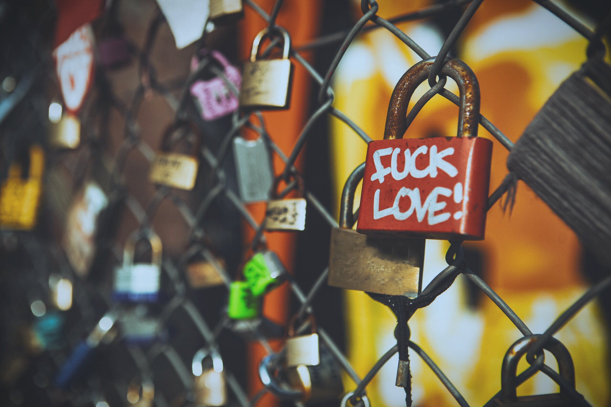 Close-up photograph of numerous padlocks attached to a chain-link fence, with a red lock in sharp focus bearing the handwritten message 'FUCK LOVE!' in white paint. The surrounding locks are blurred by a shallow depth of field, while warm orange and yellow tones in the background create a soft, out-of-focus glow. The featured lock shows signs of rust and wear, contrasting with the romantic 'love lock' tradition typically associated with such fences.
