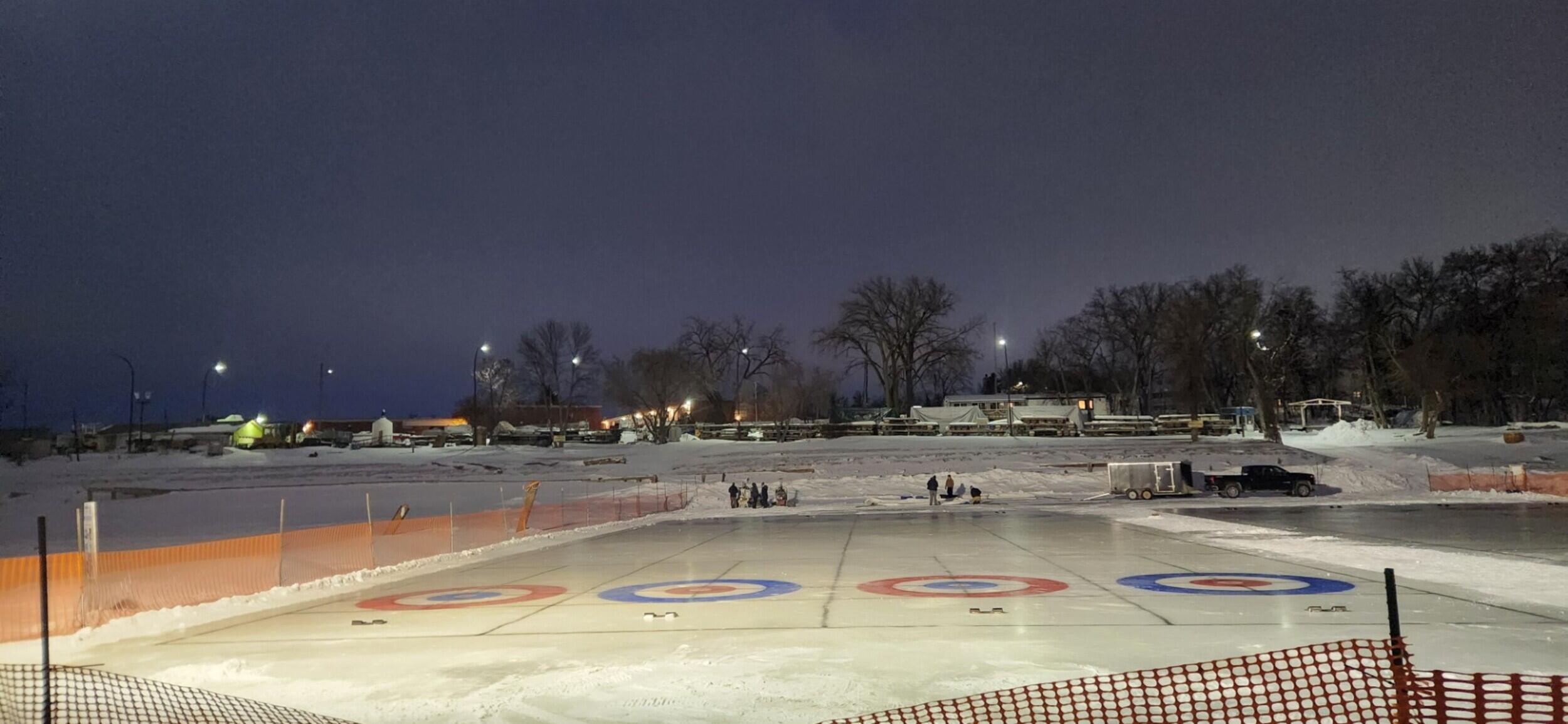 Nighttime outdoor curling rinks with orange mesh fencing. The ice features painted curling rings with blue and red circles. In the background, a group of people stands near a trailer and a pickup truck in a snow-covered field under a dark sky.