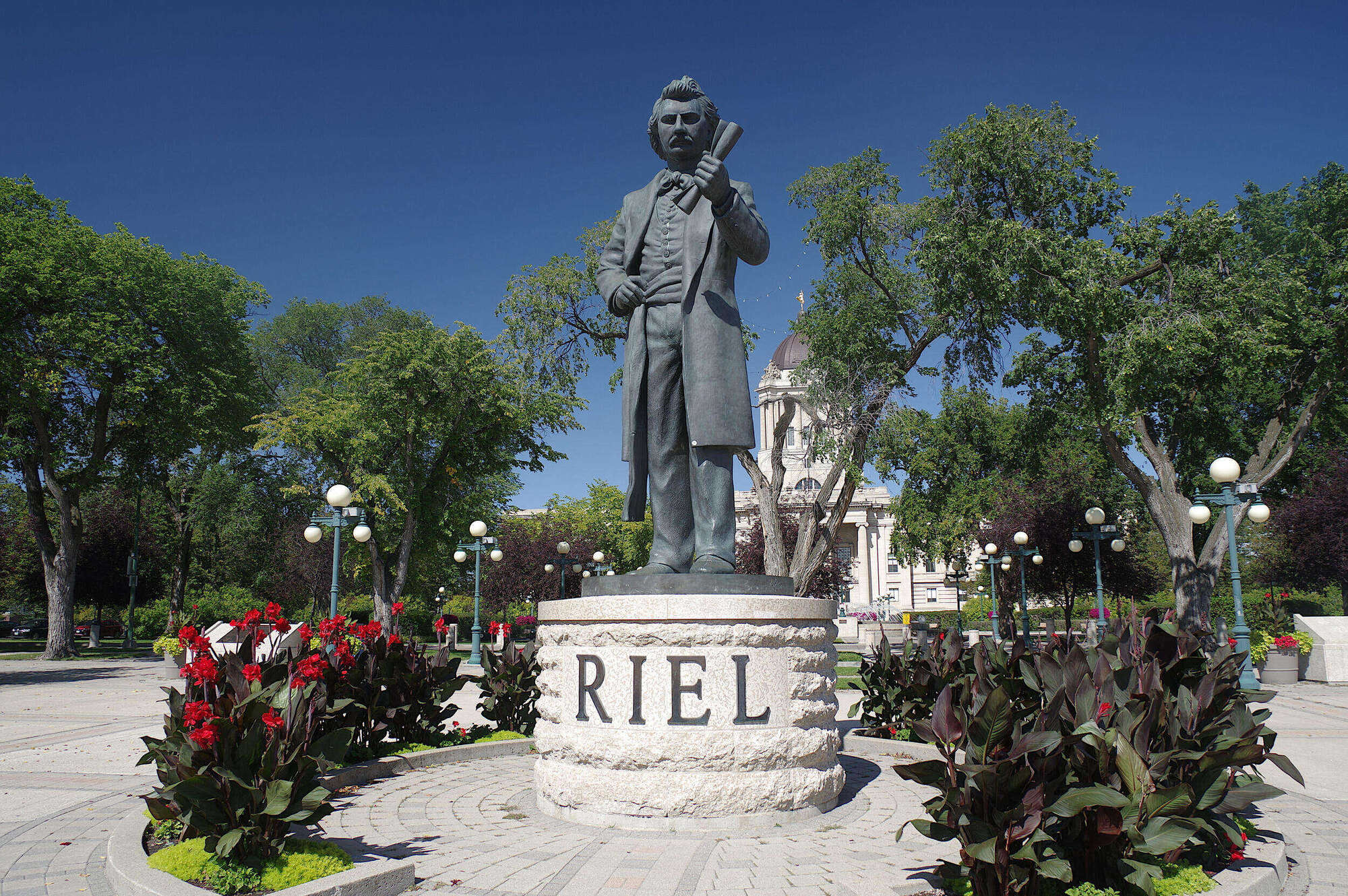 A large bronze-colored statue of Louis Riel standing on a rough-hewn stone pedestal carved with the word 'RIEL'. The statue is situated in a park-like setting with blooming red flowers, green trees, vintage lampposts, and a domed legislative building in the background under a clear blue sky.