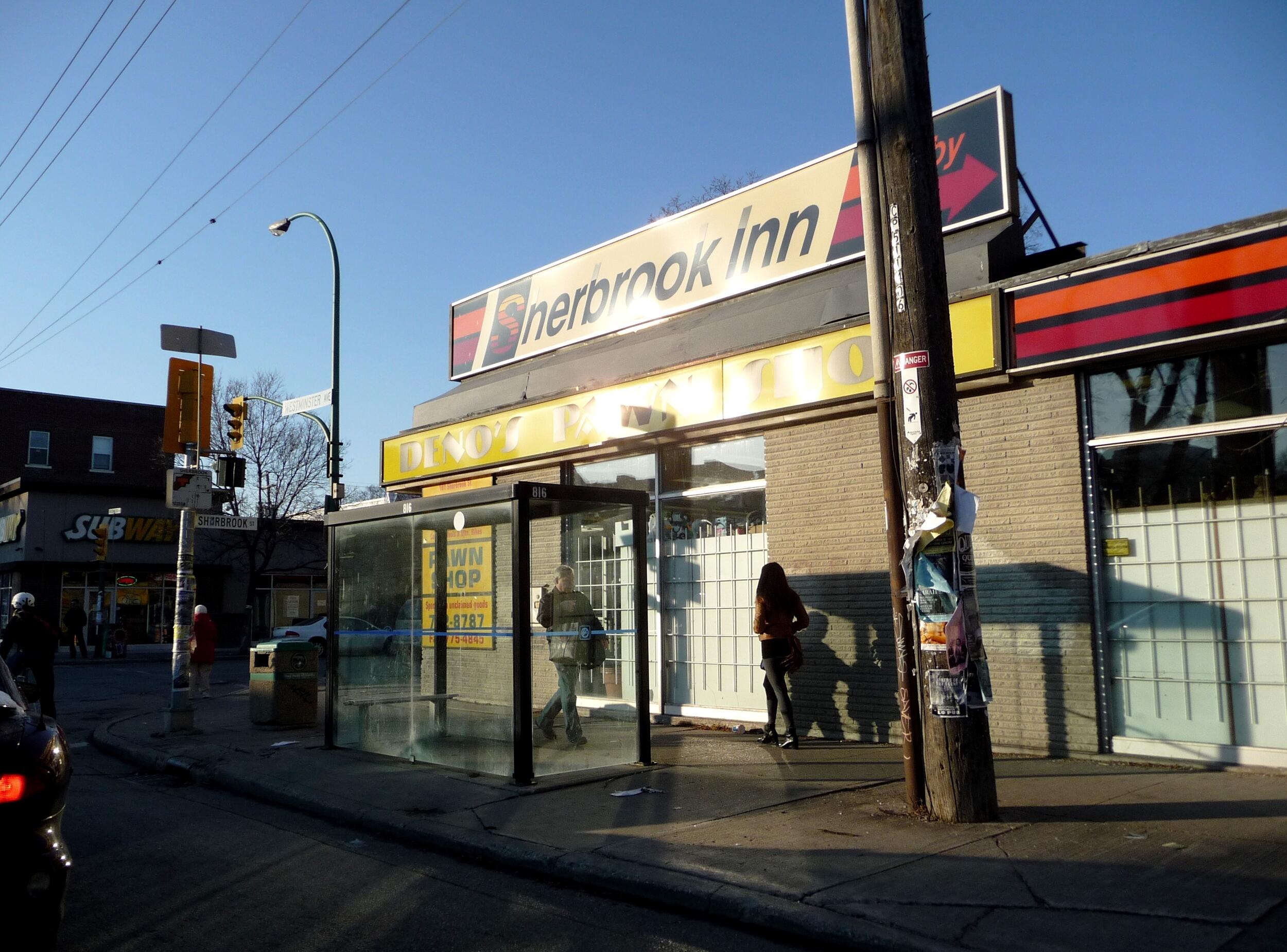 An urban street corner featuring a building with a large sign reading 'Sherbrook Inn' and a yellow awning that says 'DINER'. In front of the building is a glass bus shelter with pedestrians, and a tall wooden utility pole. A 'SUBWAY' sign is visible in the background on the left.