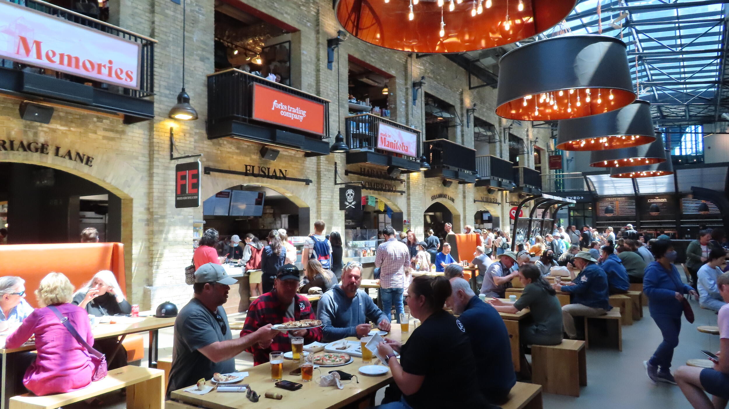 A busy, rustic indoor food hall. People are seated at communal wooden tables enjoying food and drinks. The space features brick walls, exposed steel beams, and large, distinctive cylindrical pendant lights hanging from the ceiling. Signs for 'Memories', 'Forks Trading Company', and 'Manitoba' are visible on the walls.