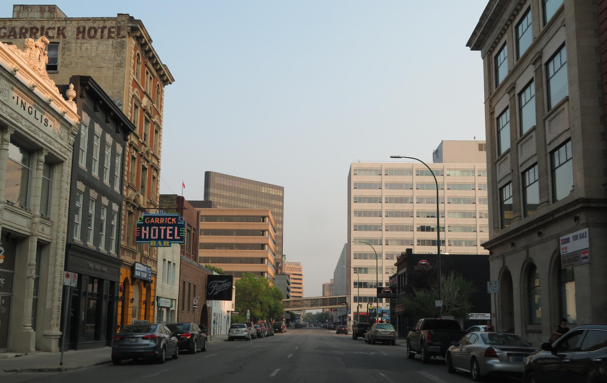 A downtown city street flanked by historic and modern architecture. On the left, a vintage brick building houses the 'GARRICK HOTEL' with a brightly lit neon sign, next to the ornate 'INGLIS' building. Cars are parked along the street, and modern office buildings and an elevated pedestrian bridge are visible in the distance.