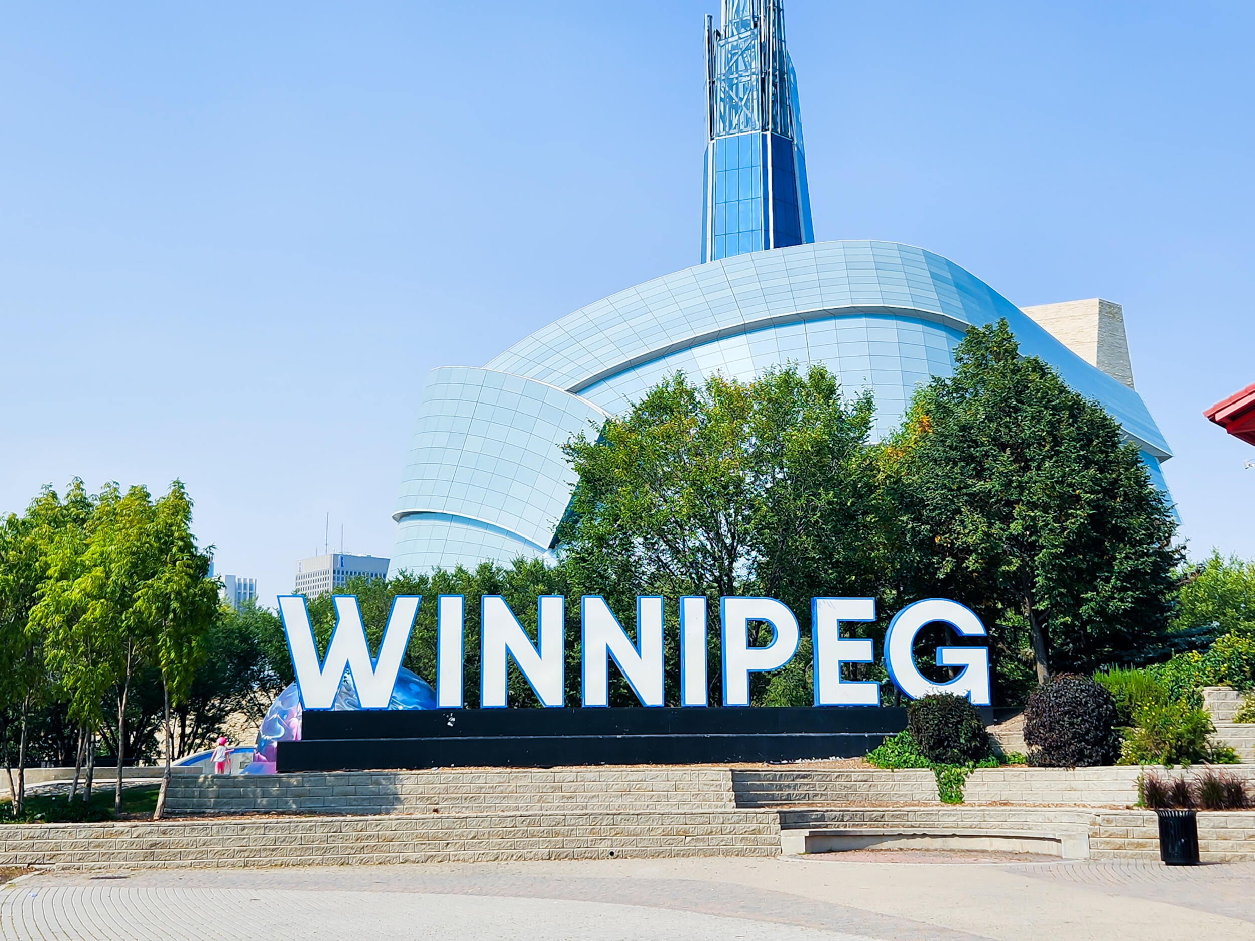 Large, white, block lettering spelling out 'WINNIPEG' mounted on a low retaining wall with tiered stone landscaping. Behind the sign, a uniquely shaped, modern blue-glass building (the Canadian Museum for Human Rights) rises prominently with a tall, glass-and-steel spire. Green trees surround the site on a sunny day.