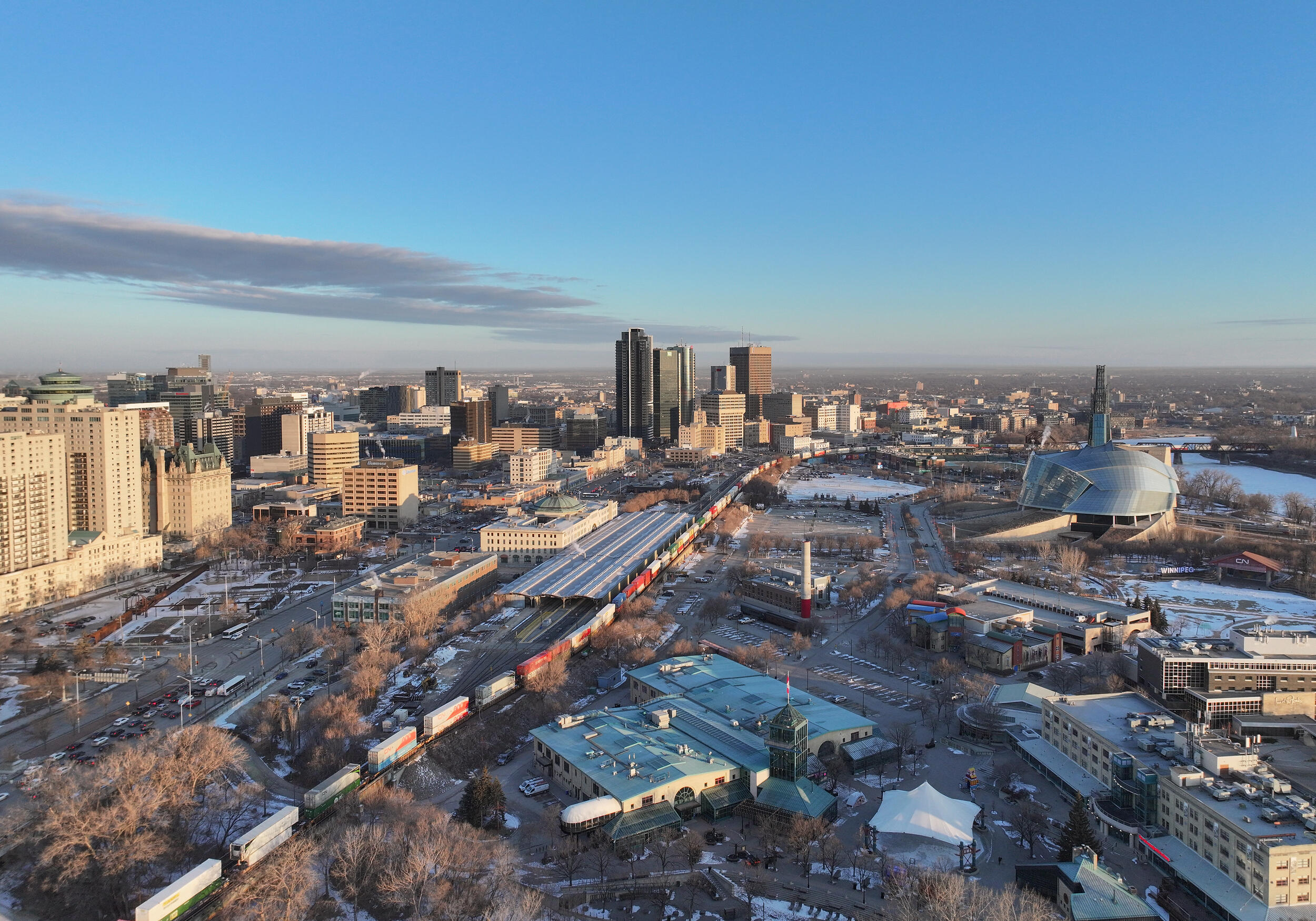 An aerial, winter view of Winnipeg. The scene includes the downtown skyline, railway tracks, a historic train station with a green roof, and the modern, sweeping glass architecture of the Canadian Museum for Human Rights. The ground is covered in snow, and the sky is clear and blue.