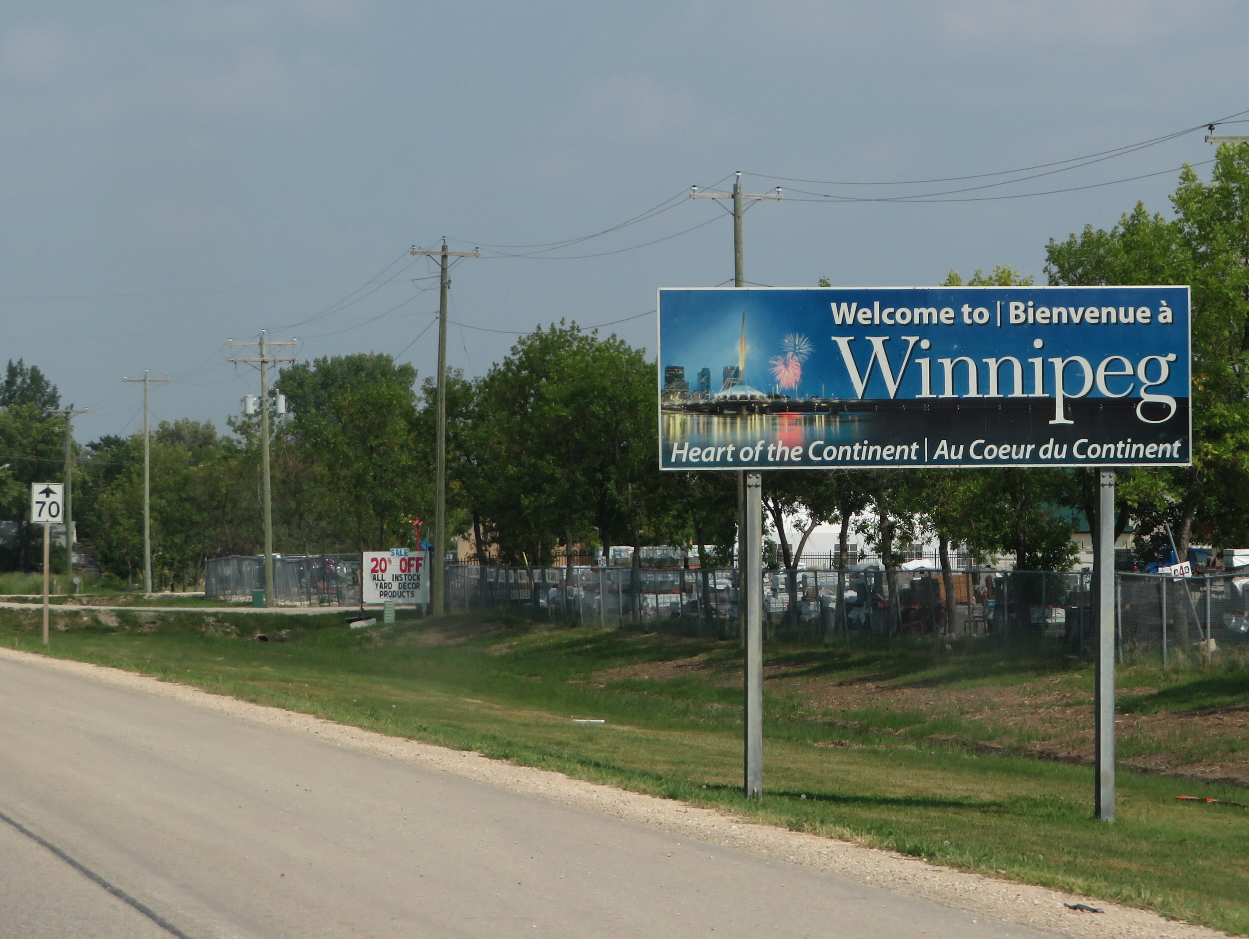 A large blue roadside billboard on a grassy verge. The sign reads 'Welcome to | Bienvenue à Winnipeg' in large white letters. Below that, it says 'Heart of the Continent | Au Coeur du Continent'. The sign features a graphic of a city skyline at night with fireworks. Power lines and a fence are visible behind the sign.