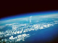 View of Earth's curved horizon from space, showing the atmosphere's blue gradient fading into black space. A rocket or space shuttle is climbing away from Earth's surface, leaving a bright white exhaust trail, with clouds scattered across the blue ocean below.