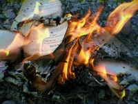 Printed pages burning on rocky ground outdoors, with tall orange flames rising from the center. The pages are from a book, and visible names include Steve Wozniak, Stanley Kubrick, and Leslie Stern, M.D., with a section header reading '[End Of Tra...' and a headline at the top beginning 'Why Did Navidson Go Back To The Hou...' The surrounding area shows ash, gravel, and dry vegetation. The image has a subtle chromatic aberration effect, giving the edges a yellow-green fringe.