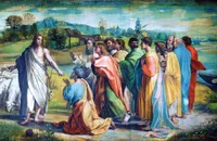 A robed Christ stands at left, gesturing toward a group of apostles in a lush pastoral landscape. At centre, an older bearded man—Peter—kneels in supplication before him, arms raised. Behind Peter, a crowd of apostles in vibrant draped robes of coral, green, gold, blue, and red look on with varying expressions of attention and concern. A flock of white sheep grazes at left. In the background, rolling green hills, scattered trees, and the towers of a distant village recede under a pale blue sky. The painting surface shows visible cracking consistent with age.