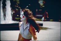 Vintage-toned photo of a young woman in profile with clown-style face paint, a red circle on her cheek and a painted winking eye. Her dark hair blowing sideways in the wind. She wears a light gingham sleeveless top and stands before a plaza fountain, with a blurred figure sitting in the background.