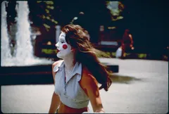 Vintage-toned photo of a young woman in profile with clown-style face paint, a red circle on her cheek and a painted winking eye. Her dark hair blowing sideways in the wind. She wears a light gingham sleeveless top and stands before a plaza fountain, with a blurred figure sitting in the background.