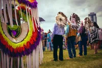 An outdoor powwow or Indigenous ceremonial gathering set against an overcast urban skyline. The left foreground is filled with a large, strikingly colourful regalia bustle. Concentric rings of neon yellow, red, orange, and purple feathers with long white ribbon fringe. In the mid-ground, several figures stand on a grassy field; two wear large traditional eagle feather headdresses that face away from the camera, their faces obscured. One person wears a Northwest Coast-style button blanket or cape featuring black-and-white formline design. The gathering includes people in both traditional regalia and everyday clothing. A highrise tower is visible through the cloudy sky in the distance, situating this cultural gathering within a contemporary urban landscape.
