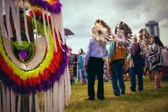 An outdoor powwow or Indigenous ceremonial gathering set against an overcast urban skyline. The left foreground is filled with a large, strikingly colourful regalia bustle. Concentric rings of neon yellow, red, orange, and purple feathers with long white ribbon fringe. In the mid-ground, several figures stand on a grassy field; two wear large traditional eagle feather headdresses that face away from the camera, their faces obscured. One person wears a Northwest Coast-style button blanket or cape featuring black-and-white formline design. The gathering includes people in both traditional regalia and everyday clothing. A highrise tower is visible through the cloudy sky in the distance, situating this cultural gathering within a contemporary urban landscape.