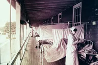 A colorized historical photograph of an open-air hospital ward, likely from the 1918 influenza pandemic. Rows of metal-framed beds with white canopy curtains line a long covered porch with wooden floors and large windows overlooking trees. A nurse in a white uniform, cap, and face mask leans over a patient in the foreground; a second figure in white stands further down the ward. A small bedside table holds medical supplies.