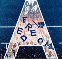Aerial photo of a triangular street mural spelling FREEDOM in navy blue letters, painted on a road or court marked with white lines and scattered with fallen autumn leaves. The letters hang like marionettes from strings held by pale puppeteer hands at the top, while a dark-skinned hand holds scissors poised to cut the strings. Faded background text reads words like HATE, FEAR, LIES, DOUBT, MURDER, DECEPTION, and PRIDE. Rows of raised human hands in varied skin tones reach up from the bottom of the triangle toward the letters. The mural is signed 'G. Johnson' in the bottom right corner.