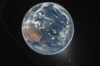 A photograph of Earth from deep space, slightly off-centre and partially in shadow, with a thin luminous blue atmospheric halo visible along the sunlit edge. Australia's arid interior is visible as a rust-coloured landmass on the left side. Swirling cloud systems cover much of the Pacific Ocean to the right. Stars are faintly visible in the black background, and a bright lens flare appears near the lower right.