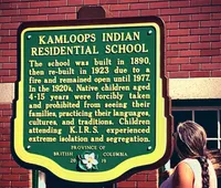 A green historical plaque mounted on a brick wall titled 'Kamloops Indian Residential School,' describing the school's operation from 1890 to 1977 and the forced removal, isolation, and cultural suppression of Indigenous children; a person stands partially visible to the right, looking at the sign