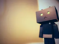 A cardboard box robot figure (Danbo-style) with round glowing yellow eyes and a triangular mouth, standing against a warm, softly blurred beige and cream background. The figure is positioned on the right side of the frame, gazing off to the left, with the left portion of the image left empty and softly lit.