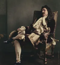 A hand-colored photograph of a male stage actor in 17th-century French theatrical costume, reclining languidly in an ornate gilded armchair. He wears a deep burgundy velvet coat with elaborate lace cravat and cuffs, white stockings with gold embroidered detail, and black heeled shoes.