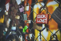 Close-up photograph of numerous padlocks attached to a chain-link fence, with a red lock in sharp focus bearing the handwritten message 'FUCK LOVE!' in white paint. The surrounding locks are blurred by a shallow depth of field, while warm orange and yellow tones in the background create a soft, out-of-focus glow. The featured lock shows signs of rust and wear, contrasting with the romantic 'love lock' tradition typically associated with such fences.