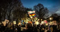 A crowd of protesters gathered at dusk holding signs and waving transgender pride and rainbow flags. Visible signs read 'LOVE TRUMPS HATE,' 'TRANS RIGHTS,' 'SAD!,' and 'NO HATE.' Bare trees and illuminated street lights frame the scene against a blue evening sky, with buildings visible in the background.
