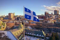 Le drapeau du Québec, le Fleurdelisé, bleu et blanc orné de quatre fleurs de lys, flotte au vent sur un mât au premier plan. En arrière-plan s'étend une vue aérienne de la ville de Québec en hiver : toits enneigés, architecture patrimoniale en briques rouges, toitures en cuivre vert aux pignons caractéristiques du Vieux-Québec, et la silhouette du centre-ville moderne à l'horizon, sous un ciel bleu parsemé de nuages dorés par la lumière rasante.