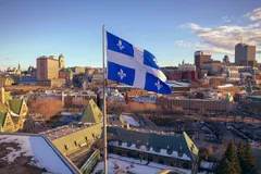 Le drapeau du Québec, le Fleurdelisé, bleu et blanc orné de quatre fleurs de lys, flotte au vent sur un mât au premier plan. En arrière-plan s'étend une vue aérienne de la ville de Québec en hiver : toits enneigés, architecture patrimoniale en briques rouges, toitures en cuivre vert aux pignons caractéristiques du Vieux-Québec, et la silhouette du centre-ville moderne à l'horizon, sous un ciel bleu parsemé de nuages dorés par la lumière rasante.