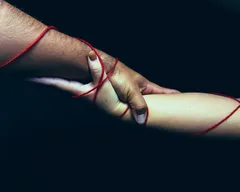 Two arms, one with darker skin, one with lighter skin, clasp together against a near-black background, bound by a single red thread that wraps around both. Dramatic low-key lighting isolates the hands at the centre, the red yarn the only point of vivid colour.