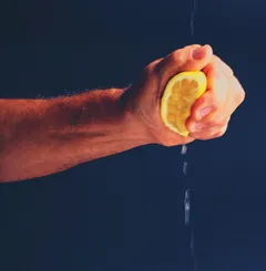 A forearm and clenched fist emerge from the left against a near-black background, squeezing a lemon half. Warm, single-source light catches the skin and the lemon's flesh; two thin streams of juice drip downward into darkness.