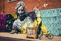 Une grande fresque murale sur un mur de briques représentant une figure de statue gréco-romaine classique rendue en noir, blanc et jaune, le regard baissé, tenant une pomme rouge à bout de bras. Des flèches et des livres ouverts flottent autour de la figure. Les sections de mur environnantes présentent des motifs décoratifs au pochoir : des fougères orange sur des fonds mauves et noirs à gauche, et de sombres motifs floraux et flocons de neige sur un fond bleu pâle à droite. Au premier plan, deux personnes sont assises sur le trottoir sous la fresque, une caisse en bois peinte d'un motif représentant une créature ailée et un véhicule se trouve à proximité, et une bicyclette est appuyée contre le mur. La fresque principale est signée « mateo » dans le coin inférieur gauche.