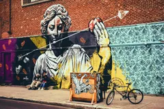 Une grande fresque murale sur un mur de briques représentant une figure de statue gréco-romaine classique rendue en noir, blanc et jaune, le regard baissé, tenant une pomme rouge à bout de bras. Des flèches et des livres ouverts flottent autour de la figure. Les sections de mur environnantes présentent des motifs décoratifs au pochoir : des fougères orange sur des fonds mauves et noirs à gauche, et de sombres motifs floraux et flocons de neige sur un fond bleu pâle à droite. Au premier plan, deux personnes sont assises sur le trottoir sous la fresque, une caisse en bois peinte d'un motif représentant une créature ailée et un véhicule se trouve à proximité, et une bicyclette est appuyée contre le mur. La fresque principale est signée « mateo » dans le coin inférieur gauche.