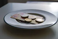 A white ceramic plate with a thin blue rim holds several euro coins of different denominations and a folded 10 euro banknote, arranged on a dark gray table or countertop with a blurred background.
