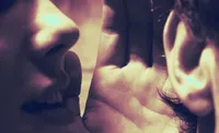Extreme close-up, grainy film photo in warm sepia-and-purple duotone: two faces nearly touching nose-to-nose, with an open palm and curled fingers resting between them in soft focus. The framing and tight depth of field create an intimate, hushed mood.