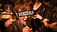 An edited photograph tinted deep orange-sepia. A person speaks forcefully into a handheld microphone while two University of Manitoba Institutional Safety Officers grip her from both sides. A thick painted banner across her eyes reads 'DENIER' in bold block letters.